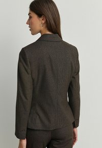 Blazer à carreaux marron avec un design structuré, coupe ajustée, col à revers et poignets boutonnés. Tissu texturé avec des détails de motif subtils.
