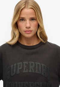 Sudadera de cuello redondo en gris oscuro con letras en relieve "SUPERDRY UNIVERSITY", confeccionada en suave material de algodón con un acabado ligeramente texturizado.