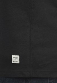 Sort bomulds-T-shirt med en synlig søm, med et lille hvidt mærke syet nederst til venstre, der står "BLEND GLOBAL JEANMAKER BUILT TO BLUE."