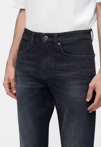 Homme portant un jean noir délavé et une chemise blanche texturée à manches courtes, les mains détendues le long du corps.