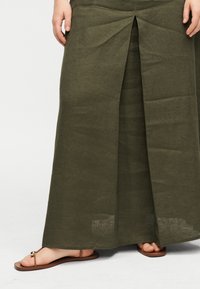 Κάτω άκρα που φοράνε μια μακριά φούστα σε ελαιοπράσινο χρώμα με άνοιγμα μπροστά και καφέ επίπεδα σανδάλια με δαχτυλίδι για τα δάχτυλα και μεταλλικές χάντρες.