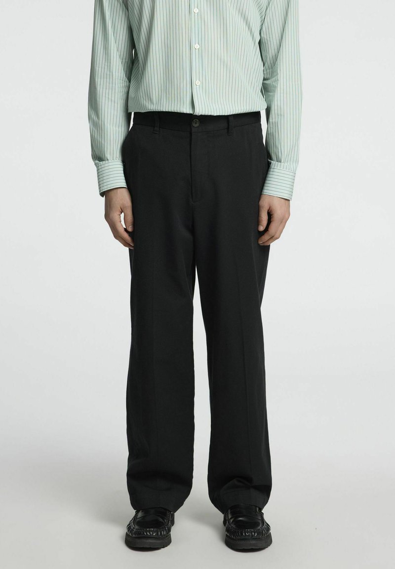 Juodos kelnės tiesaus kirpimo, derinamos su žaliai dryžuota ilgomis rankovėmis marškiniais. Atrodo, kad medžiaga yra audinė, su lygiu paviršiumi.