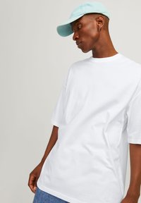 Bílá oversized bavlněná tričko s krátkým rukávem, kulatým výstřihem a volným střihem. Nosené s světle modrou baseballovou čepicí.