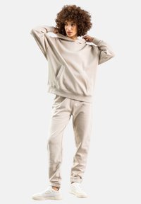 Beige sweatshirt med hætte og frontlomme, parret med matchende tapered sweatpants. Sættet er lavet af blødt stof; modellen har hvide sneakers på.