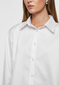 Hvid skjorte med knapper og klassisk krave, der har perlelignende knapper og blød, glat stoftekstur. Nærbillede af halsudskæring.