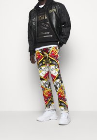 Black quilted bomber jacket over a black hoodie with gold text, paired with patterned pants in red, white, and yellow. White sneakers.