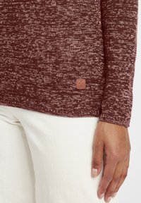 Maroon textured sweater with a rounded hem and long sleeves. Features a small brown logo patch near the hem. Paired with white pants.