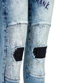 Jean en denim bleu clair avec des patchs à paillettes argentées et des trous effilochés au niveau des genoux laissant apparaître un tissu noir dessous.