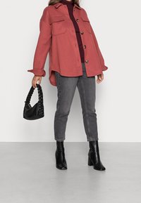 Kobieta w różowej zapinanej kurtce, burgundowym sweterku z golfem, szarych dżinsach, czarnych botkach na obcasie, trzymająca czarną torebkę z plecionym uchwytem przy sobie.