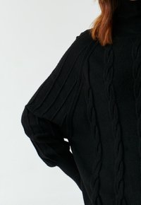 Pull en tricot noir avec un col montant, présentant un motif en câble texturé et des manches amples. Tissu doux et chaud.