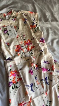Abito floreale in tessuto beige chiaro, caratterizzato da fiori viola, arancioni e rosa. Il design include maniche corte, chiusura frontale con bottoni e una cintura in vita.