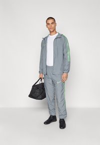 Grauener Trainingsanzug mit grünen Seitenstreifen, Reißverschlussjacke, Hose mit elastischem Bund, kombiniert mit einer schwarzen Sporttasche und schwarzen Schuhen.