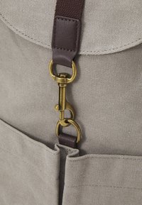 Primo piano di una borsa di tessuto beige con tracolla in pelle marrone e chiusura in ottone antico che fissa il flap anteriore alla tasca.