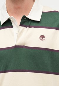 Πόλο μπλούζα με γιακά, με φαρδιές οριζόντιες ρίγες σε πράσινο, κρεμ και μωβ και μικρό κέντημα λογότυπου δέντρου στο στήθος.