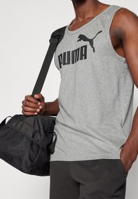 Grijze tanktop met zwart "PUMA" logo en kattensilhouet. De stof lijkt zacht; gedragen met donkere broek en een zwarte sporttas.