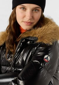 Schwarze Daunenjacke mit einem Kunstpelzkragen, Reißverschluss, glänzender Oberfläche und einem Logopatch am Ärmel. Orangefarbene Innenlage sichtbar.