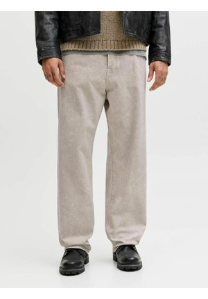 Personne portant un pantalon large gris clair, des chaussures en cuir noires, un pull en tricot marron et une veste en cuir noire, debout les mains détendues.