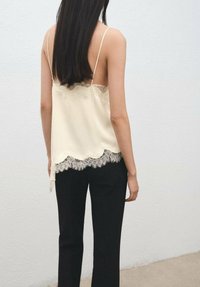 Camiseta sin mangas color crema con borde de encaje en el dobladillo, tirantes finos, combinada con pantalones negros de corte entallado. Textura suave, diseño elegante con énfasis en el encaje.