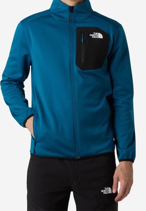 Mann trägt blaue Jacke mit Reißverschluss, schwarzer Brusttasche und schwarzer Hose, beide mit The North Face-Logo, Hände in den Taschen.