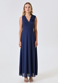 Marineblaues ärmelloses Kleid mit V-Ausschnitt, gerafftem Oberteil und floralem Akzent an einer Schulter. Fließender Rock mit leichtem Glanz.