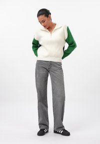 Cream knitted pullover with a quarter zip, featuring green and navy accents on the sleeves; paired with gray straight-leg jeans and black sneakers.