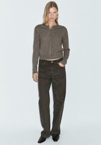 Cardigan marrone con bottoni dorati, abbinato a jeans a vita alta verde scuro. Il modello indossa scarpe nere a punta. Tessuto texturizzato e design aderente.