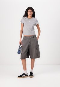 Pilkos medvilnės marškinėliai trumpomis rankovėmis su nedideliu logotipu. Palaidi pilki šortai su šoninėmis kišenėmis ir mygtuko uždarymu. Juodi sportbačiai su baltais dryžiais.