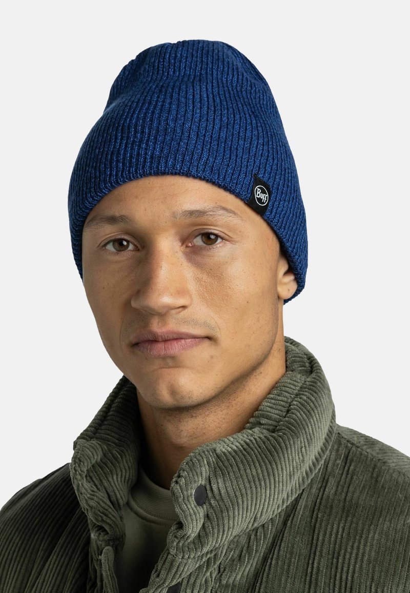 Knitted blue beanie with a ribbed texture. Logo patch on the side. Paired with a green corduroy jacket with a high collar.