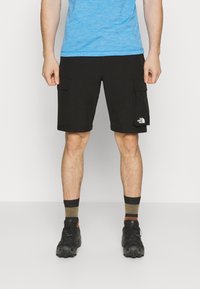 Svarta cargo shorts med sidofickor, i kombination med en blå träningsskjorta. Svarta skor och mörkrandiga strumpor syns också.