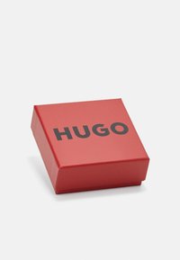 Boîte cadeau en carton rouge avec une texture lisse, avec de grandes lettres noires épelant "HUGO" sur la surface supérieure. Forme carrée avec un couvercle articulé.