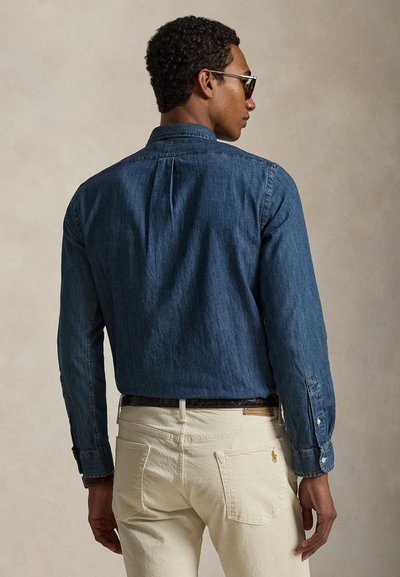 Uomo che indossa una camicia di denim blu scuro e pantaloni color crema, visto di spalle su uno sfondo beige uniforme.
