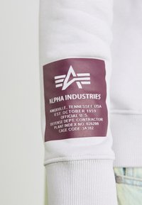 Manche de sweat-shirt blanc avec une étiquette violet foncé portant un texte blanc et un logo. L'étiquette comprend le nom de la marque et la date de création.