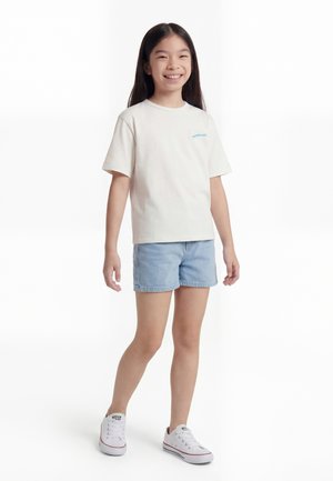 Fată tânără în picioare, zâmbitoare, purtând un tricou simplu de culoare alb-crem, pantaloni scurți de denim albastru deschis și adidași albi, pe un fundal alb.