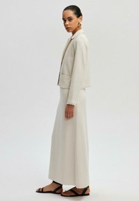 Beige linnjacka med pärlprydnader och fickor, ihopparad med en matchande lång kjol. Svarta sandaler med platta remmar och guldhårdvara.