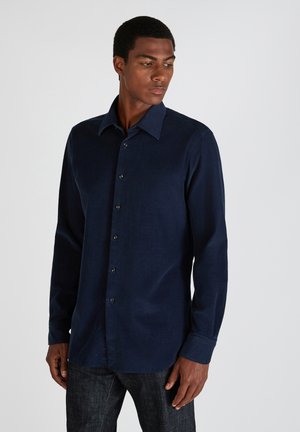 Man in een donkerblauwe, langarmige overhemden met knopen en donkere spijkerbroek, kijkend naar beneden en naar links tegen een effen achtergrond.