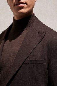 Homme portant un blazer texturé marron foncé et un pull à col roulé assorti, montré des épaules aux lèvres sur un fond uni.