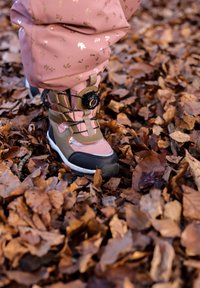 Bota de senderismo marrón y rosa con detalles en negro, superficie texturizada y sistema de cierre redondo. Colocada sobre un lecho de hojas secas marrones.