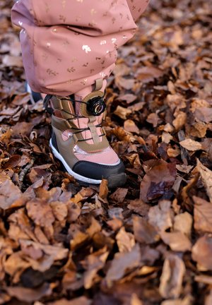 Bruine en roze wandelschoen met zwarte accenten, een gestructureerd oppervlak en een rond sluitsysteem. Geplaatst op een bed van droge, bruine bladeren.