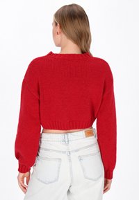 Roter, kurzer Pullover im groben Strickdesign mit geripptem Saum und weiten Ärmeln, kombiniert mit hellblauen, hochgeschnittenen Jeans.