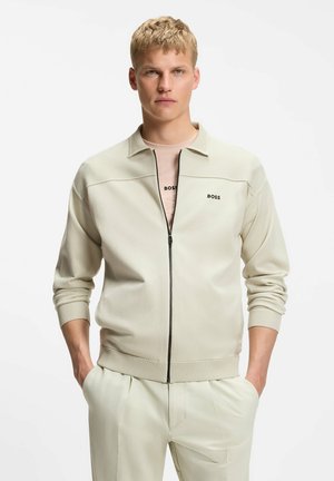 Junger Mann mit kurzen blonden Haaren, der eine beige Reißverschlussjacke mit „BOSS“-Logo und dazu passende hellfarbige Hose trägt, Hände in den Taschen.