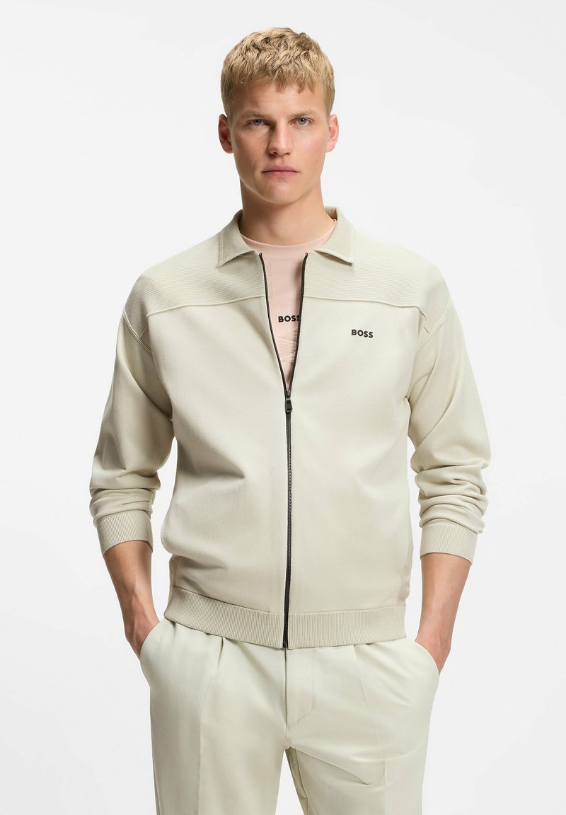 Jeune homme aux cheveux blonds courts portant une veste beige zippée avec le logo « BOSS » et un pantalon clair assorti, mains dans les poches.