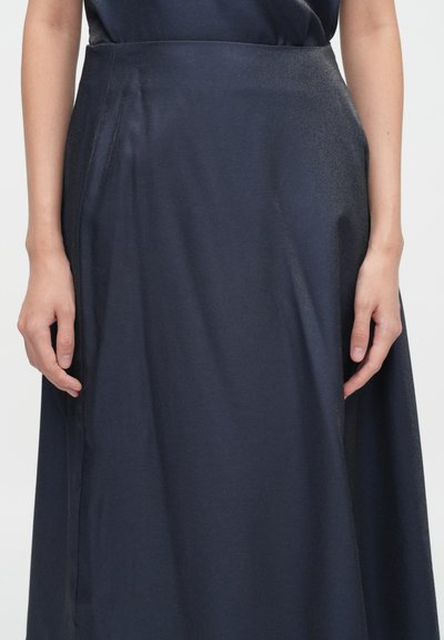 Temno modra dolga krila z gladko teksturo, stranskimi naborki in prilegajočim pasom, dosežejo do sredine tele. Roke postavljene blizu pasu.