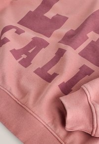 Sweat-shirt rose avec de grandes lettres épaisses rose foncé formant le mot « CALL » et avec des poignets et un ourlet côtelés visibles dans le cadre.