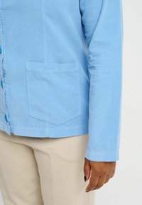 Camicia di corduroy blu chiaro con maniche lunghe, chiusura frontale a bottoni e taschino sul petto. Tessuto strutturato e dettagli di cucitura delicati.
