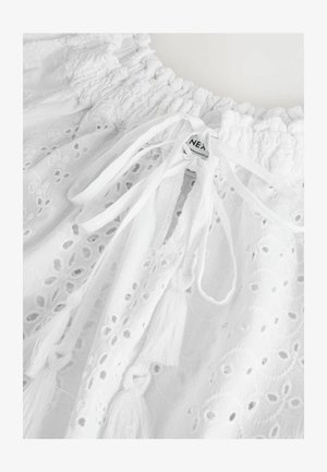 Valge puuvillane topp, millel on tikitud lillemuster, koondatud kaelus, nöörid ja tupsud ning silt "NEXT."