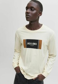 Kreemjast pikast varrukast t-särk, mille esiküljel on musta ja pruunika ristkülikukujuline logo, millel on kiri "JACK & JONES COLLECTIONS".