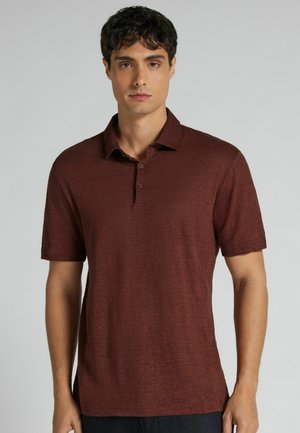 Mężczyzna o krótkich ciemnych włosach, ubrany w jednokolorową bordową koszulkę polo z krótkim rękawem, stojący na jednolitym jasnoszarym tle.