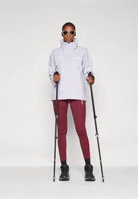 Asmuo su akiniais nuo saulės, šviesiu švarku ir bordo spalvos leggingais stovi, laikydamas juodus žygių lazdas priešais vienodą foną.