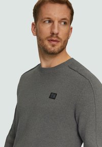 Pull en tricot gris avec un col rond, des poignets côtelés et un patch logo noir sur la poitrine. Texture lisse et design ajusté.