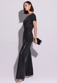 Czarny kombinezon z cekinami, krótki rękaw, dekolt w serek, pasek w talii i szerokie nogawki. Zestawiony z metalicznymi sandałami na otwartym palcu i czarną kopertówką.